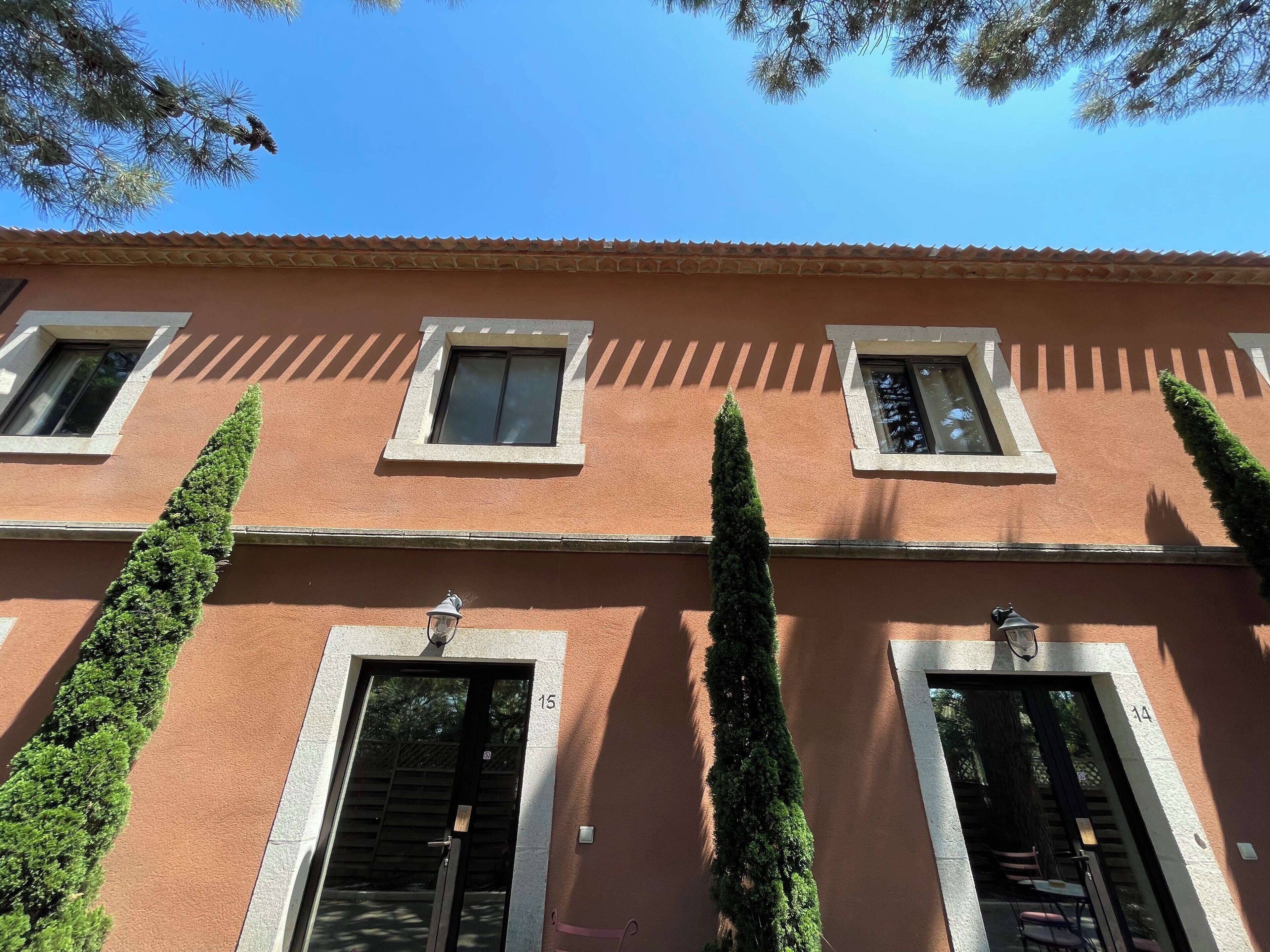 A building with three windows and two doors, each with a small window above. There are tall, narrow trees in front of the doors.