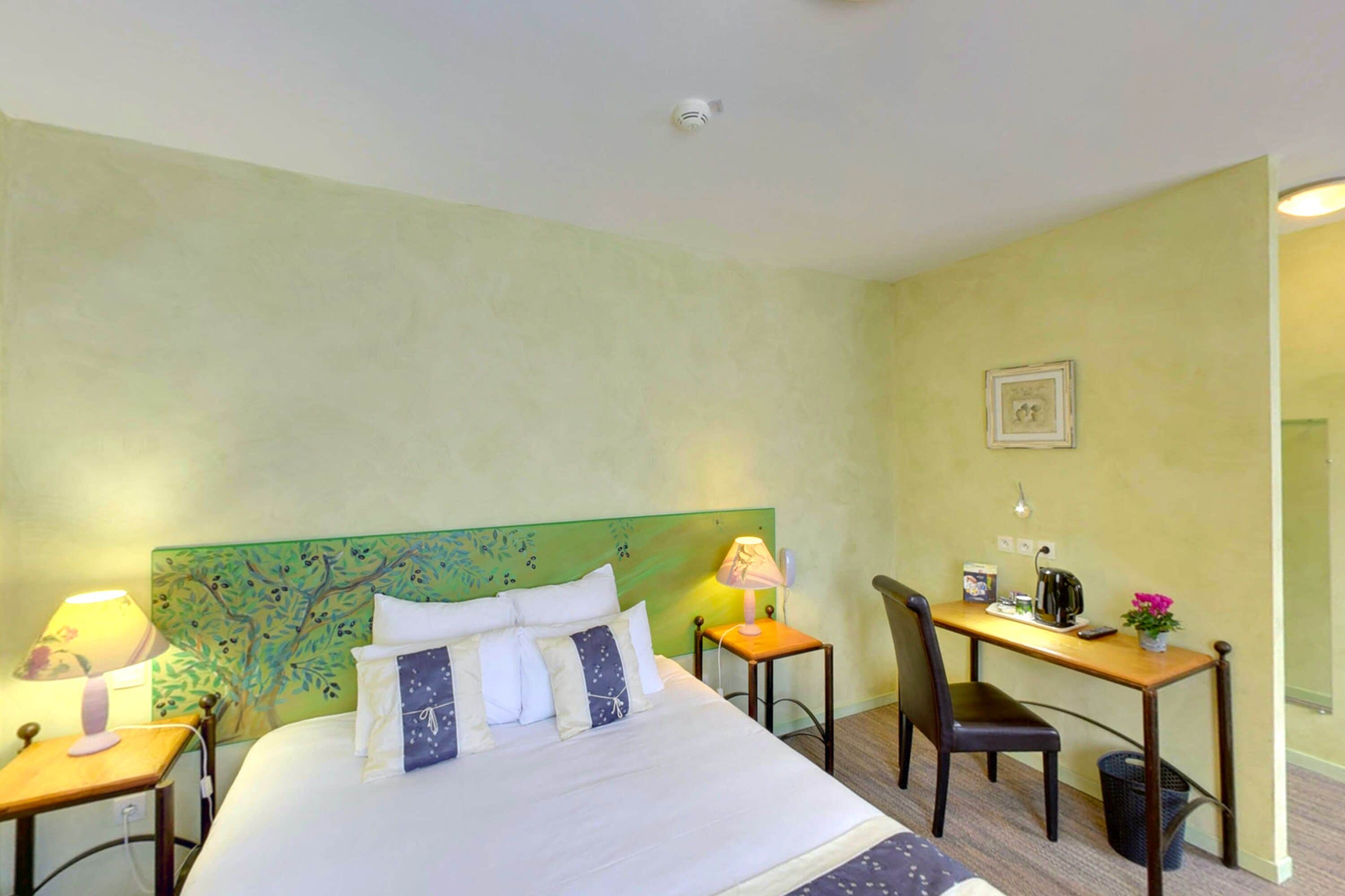 Une chambre d’hôtel avec un lit, un bureau, une chaise, une lampe et une petite table sur laquelle est posée une plante.