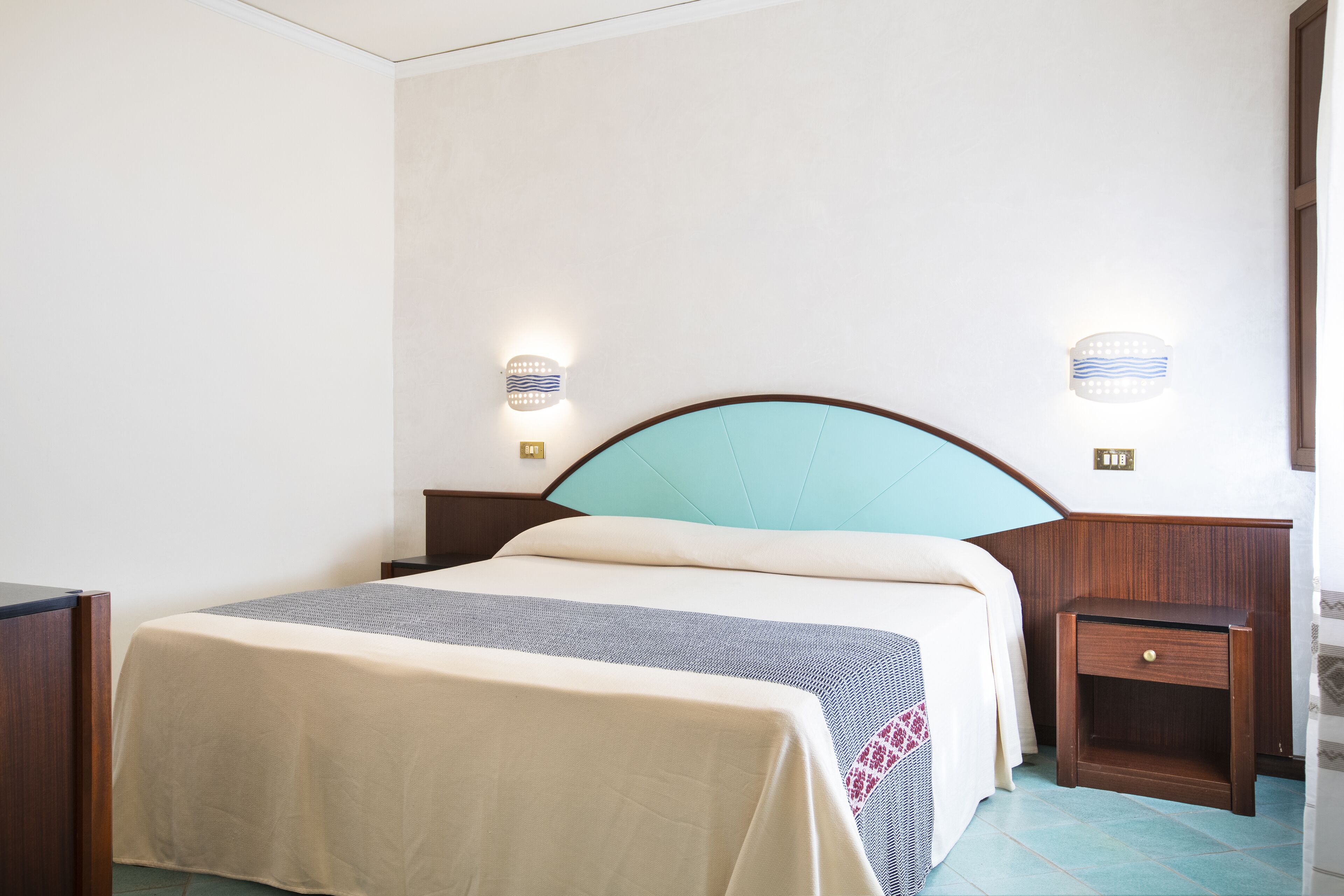 Una cama bien hecha con cabecera, mesitas de noche e iluminación en la pared.