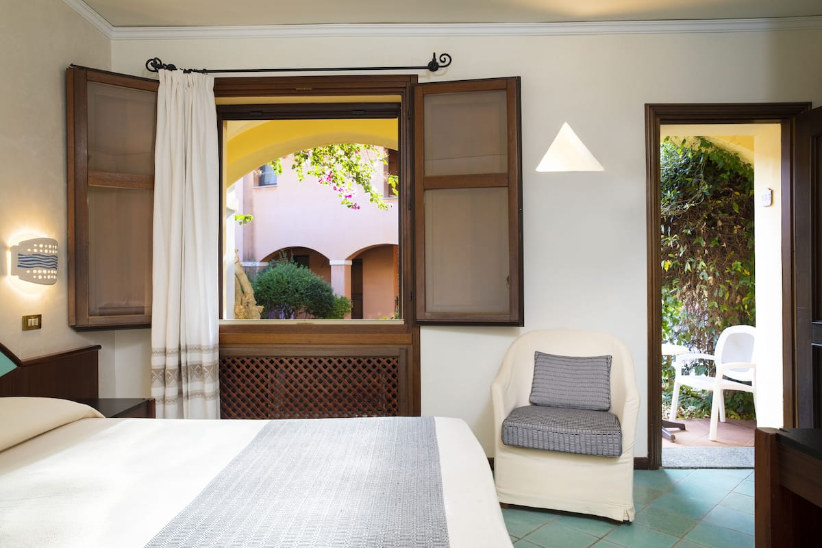 A hotel room with a bed, a chair, a window with open shutters, and a view of an outdoor courtyard.