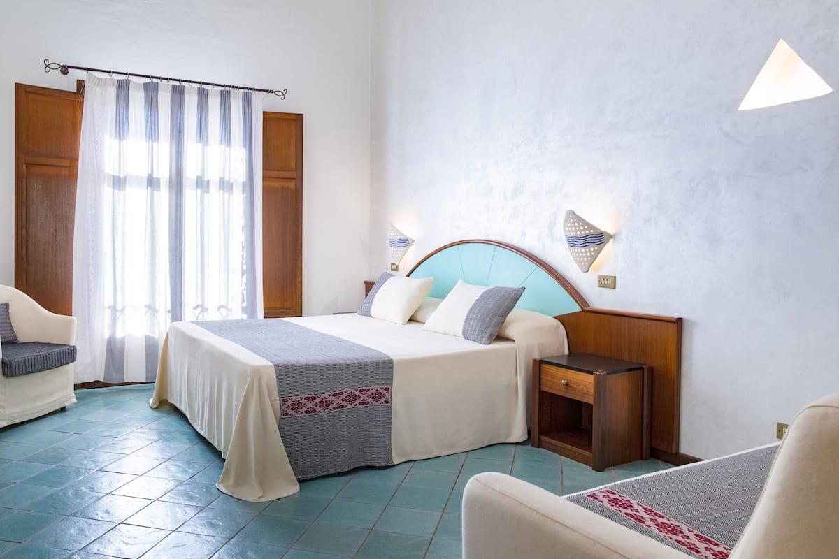 A hotel room with a bed, bedside tables, a chair, a desk, and a window with curtains.