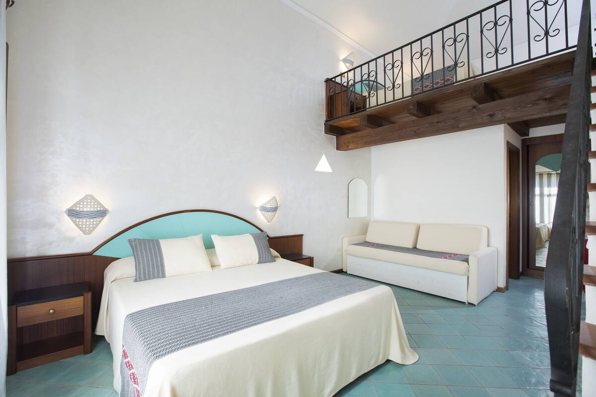 A bedroom with a bed, bedside tables, a sofa, and a balcony with a railing.