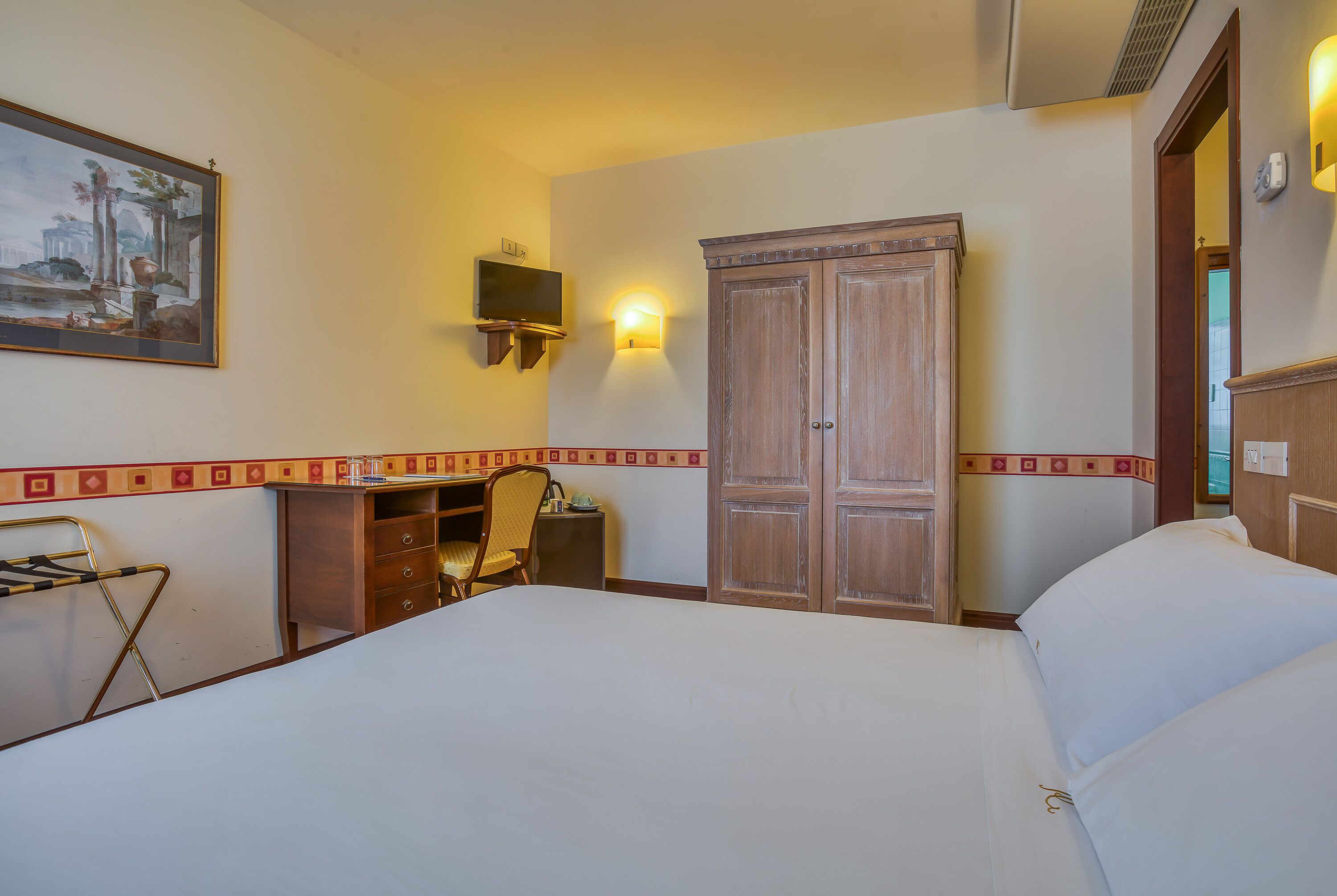 A hotel room with a bed, desk, chair, wardrobe, and a painting on the wall.