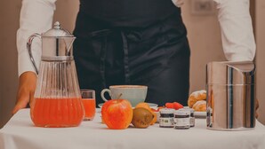 A person in a white shirt and black apron standing behind a table with a coffee pot, a glass of orange juice, an apple, and some pastries. - Hotel Des Reves - Al Poggio di Casalucense (Sant'Elia Fiumerapido)