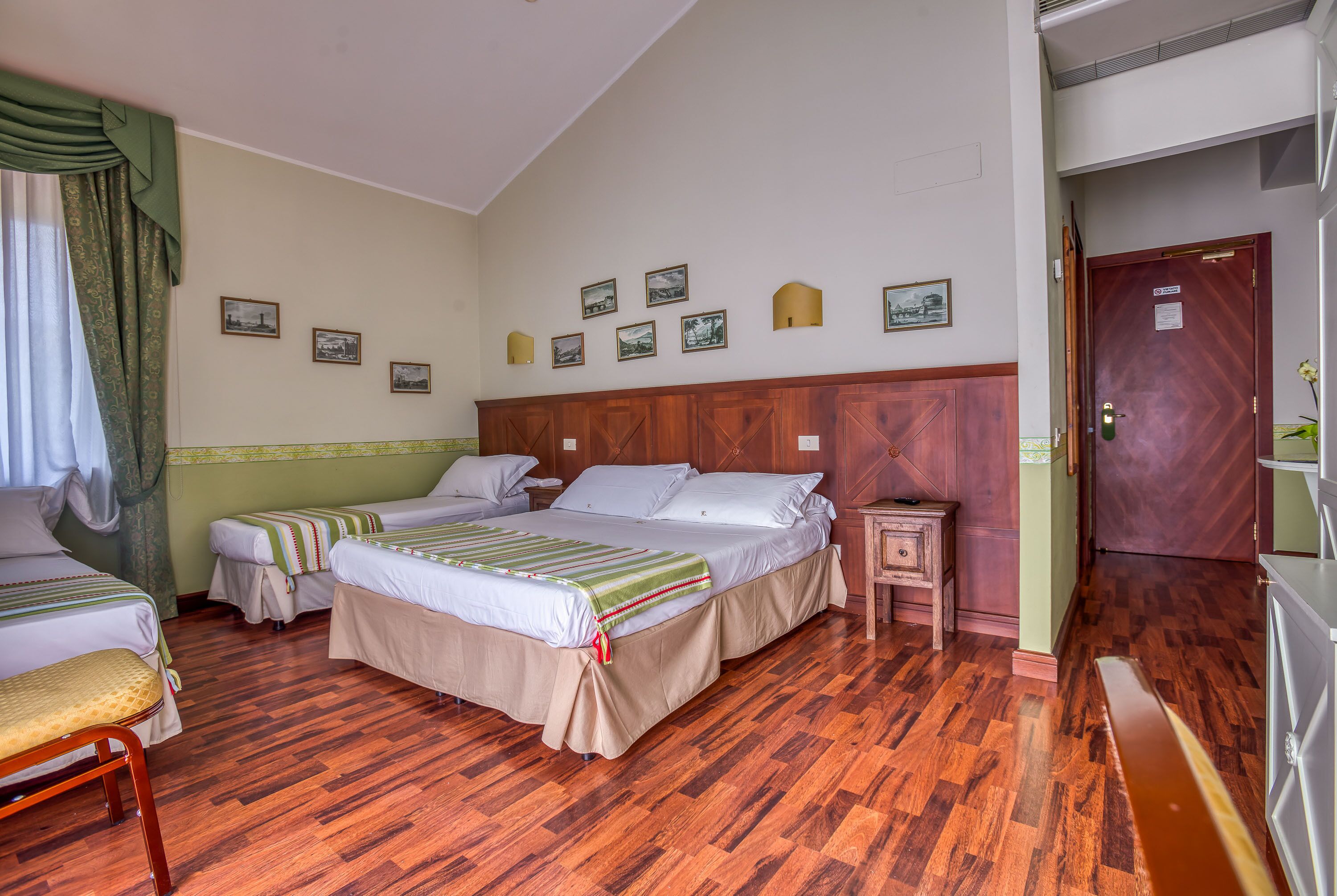A hotel room with two beds, wooden flooring, a wooden headboard, a nightstand, and a door with a sign.