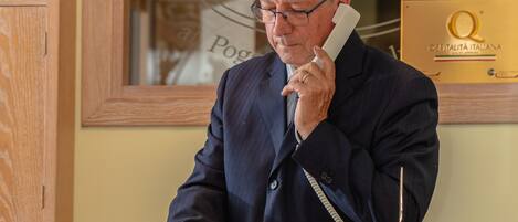 Ein Mann in einem dunklen Anzug steht an einem hölzernen Empfangstresen und hält ein Telefonhörer in der Hand.