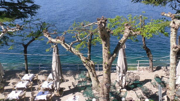 A seaside restaurant with outdoor seating, overlooking a body of water and a mountain. - Hotel Pai (Torri del Benaco)