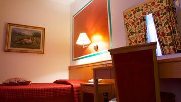 A hotel room with a bed, a desk, a lamp, a chair, a painting, and a window with curtains.