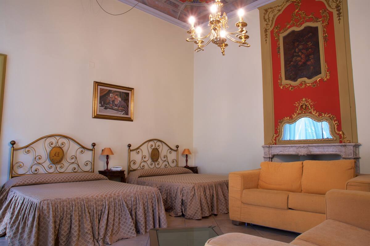 Une chambre d’hôtel avec deux lits, un canapé, un lustre, un tableau et une cheminée.