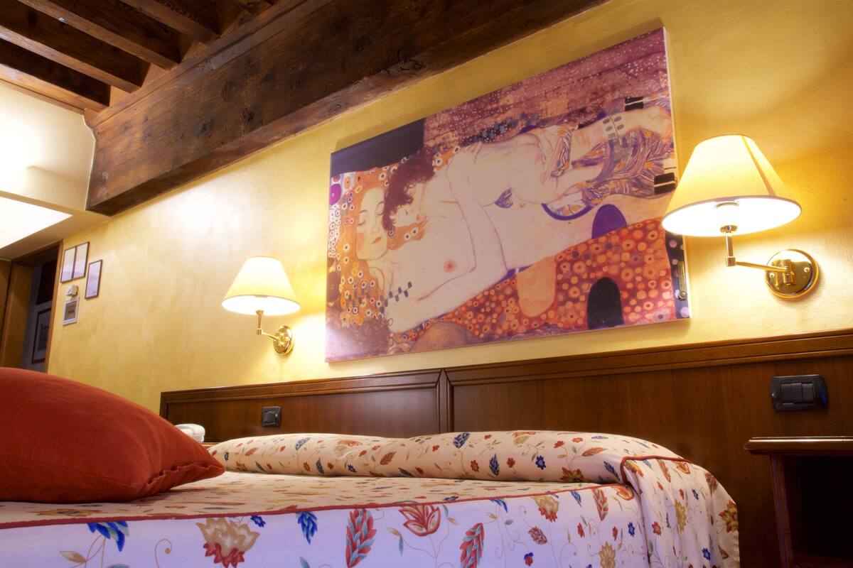 Une chambre d’hôtel avec un lit, une couvre-lit à motifs floraux, deux lampes fixées au mur et un grand tableau au-dessus du lit.