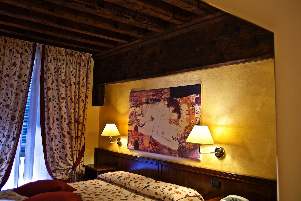 A hotel room with a wooden ceiling, floral curtains, a large painting on the wall, two bedside lamps, and a bed with floral bedding.