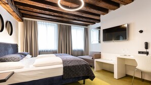 Ein modernes Hotelzimmer mit Bett, Schreibtisch, Sessel, Fernseher und einem großen Fenster mit Vorhängen.