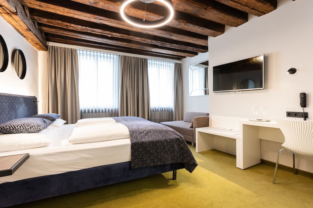 Una habitación de hotel moderna con cama, escritorio, silla, televisor y un amplio ventanal con cortinas.
