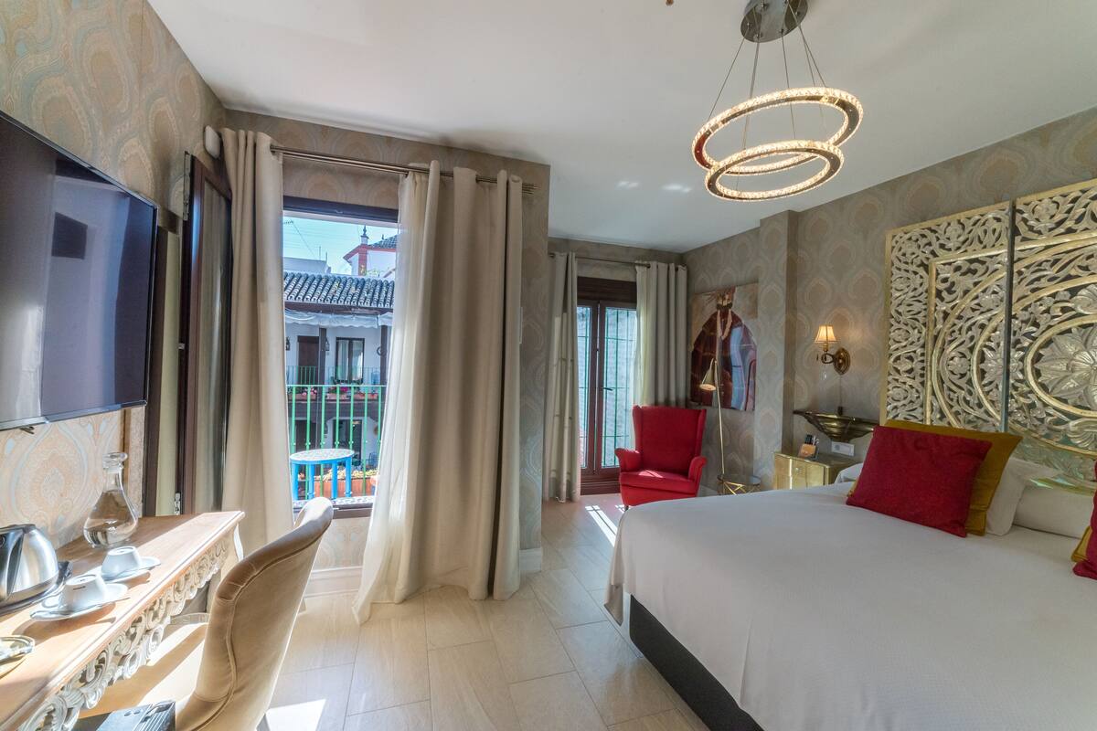 A hotel room with a bed, a TV, a desk with a chair, a red chair, a balcony with a view of buildings, and a decorative wall panel.