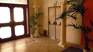 A hallway with a tiled floor, a wooden door with glass panels, a statue, and potted plants. - Hotel 4 Mori (Cagliari)
