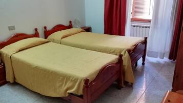 Ein Zimmer mit zwei Holzbetten, einem Nachttisch, einem Heizkörper und einem Fenster mit roten Vorhängen.