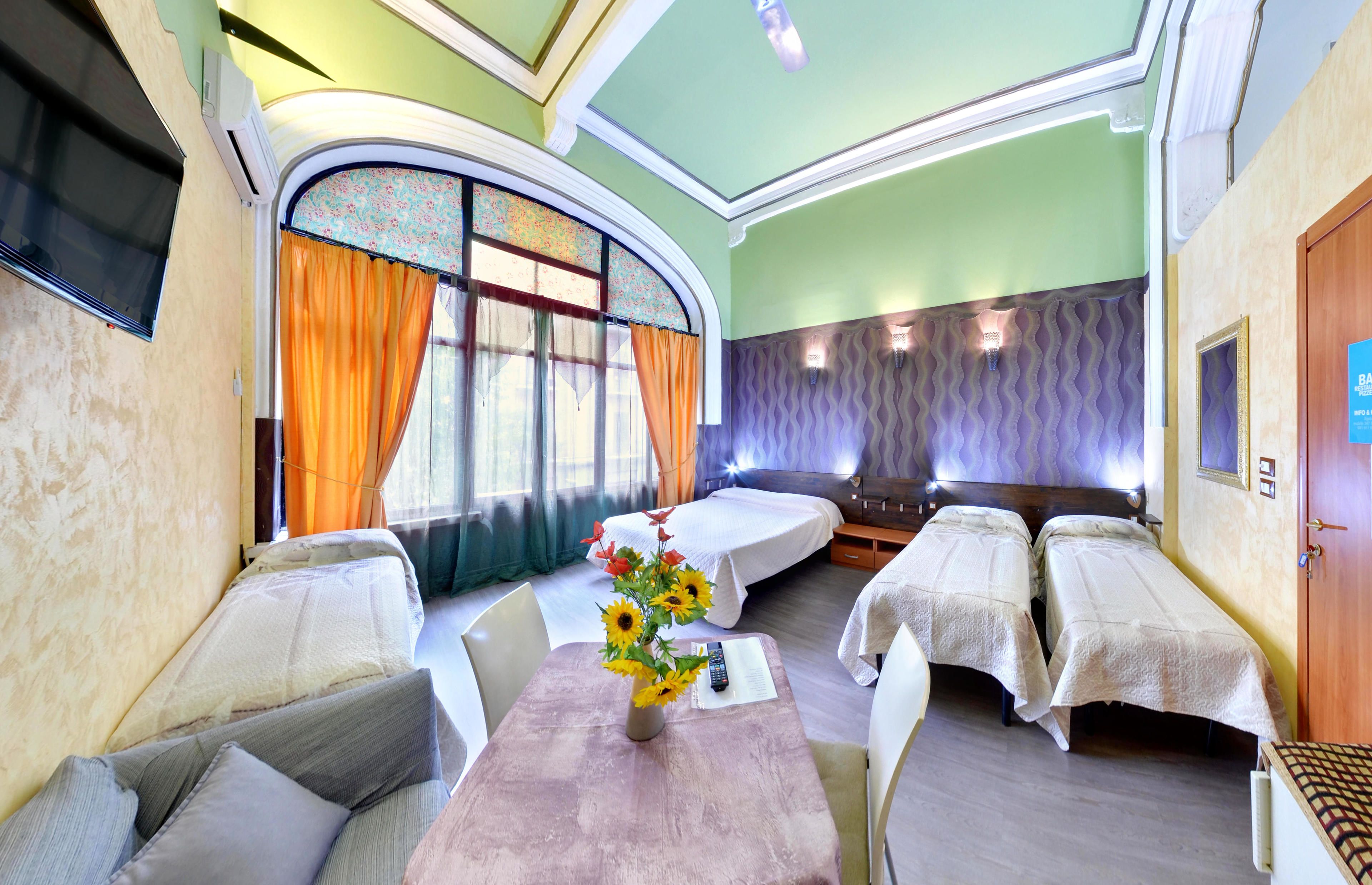 Une chambre avec deux lits simples, un canapé, une table ornée d’un vase de tournesols et une grande fenêtre avec des rideaux.