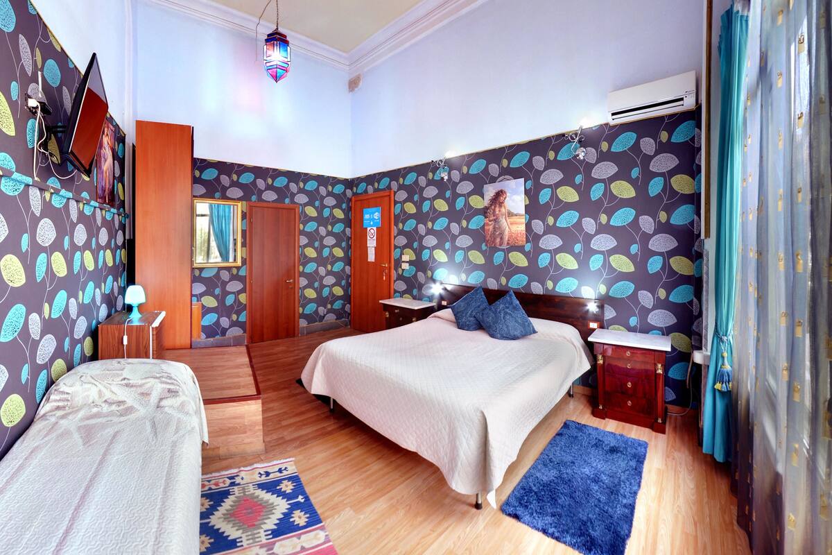 A bedroom with a bed, a bedside table, a nightstand, a lamp, a rug, and a wall with a leaf pattern.