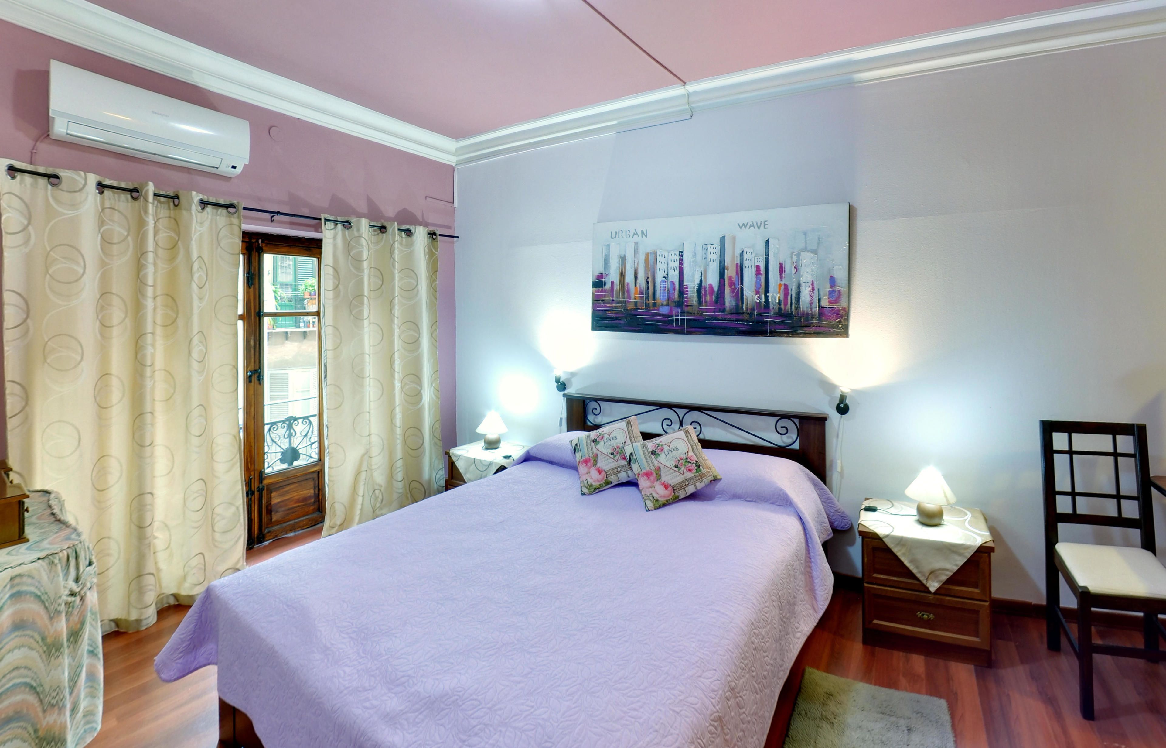 Een slaapkamer met een bed, nachtkastje, stoel, raam met gordijnen, een aan de muur bevestigde airconditioning en een stadsgezicht schilderij.