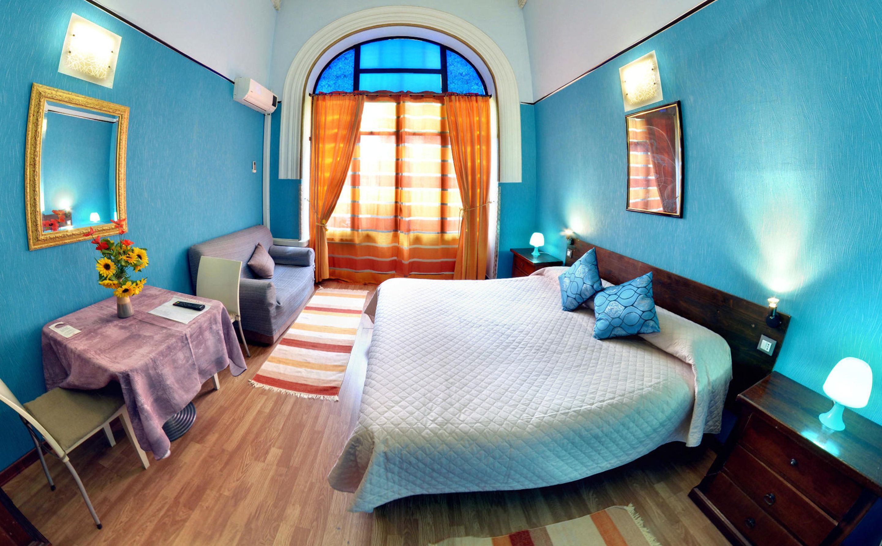 A bedroom with a bed, a sofa, a table with a vase of flowers, and a window with curtains.