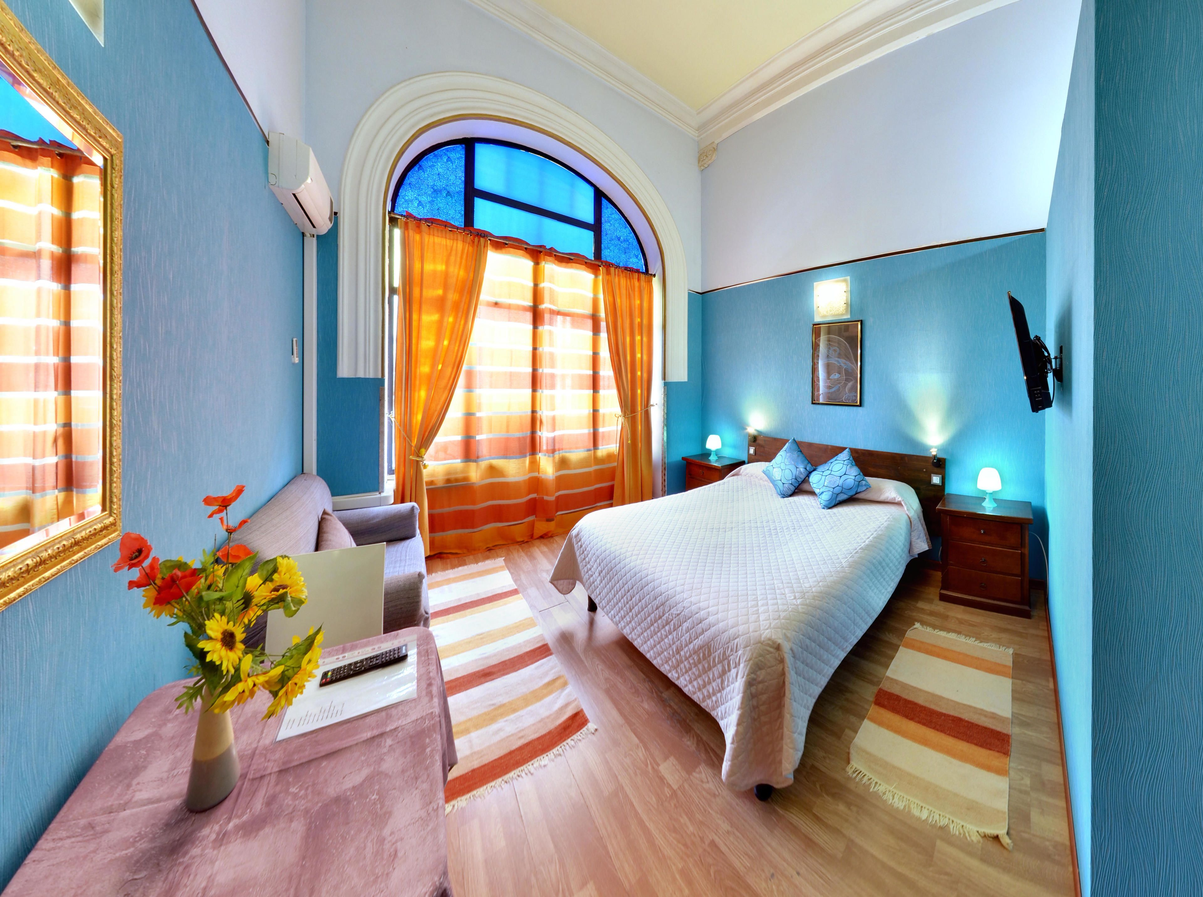 A hotel room with a bed, bedside tables, a desk with a lamp, a chair, a window with curtains, and a wall-mounted light fixture.