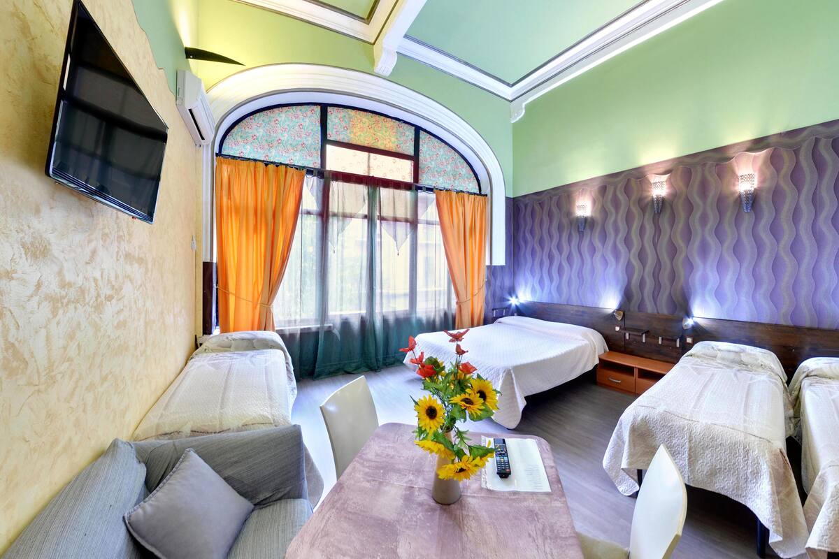 A hotel room with a sofa, a table with a vase of sunflowers, a bed with a white bedspread, and a television mounted on the wall.