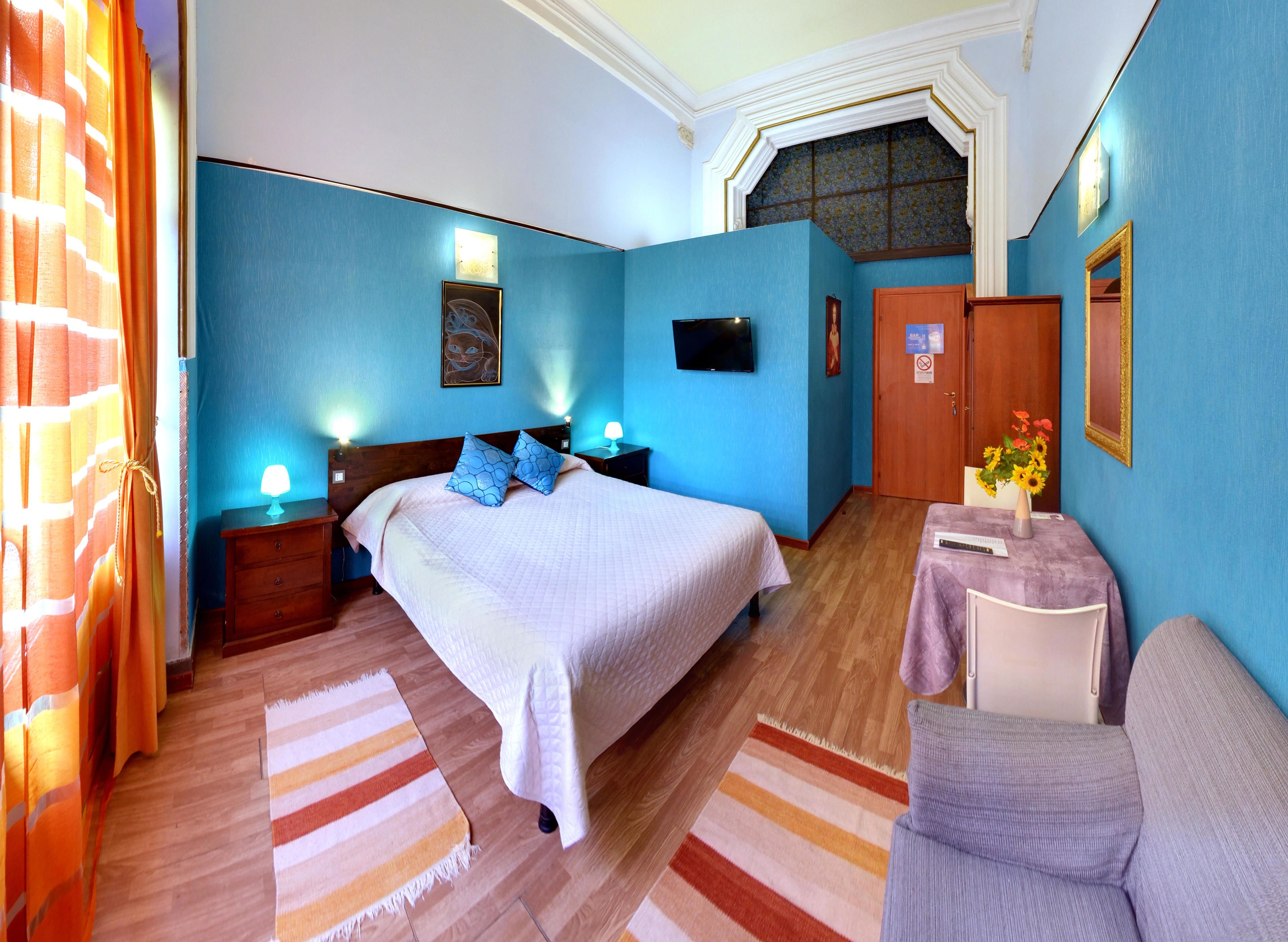 A bedroom with a large bed, bedside tables, a desk with a chair, a TV, and a door with a sign.