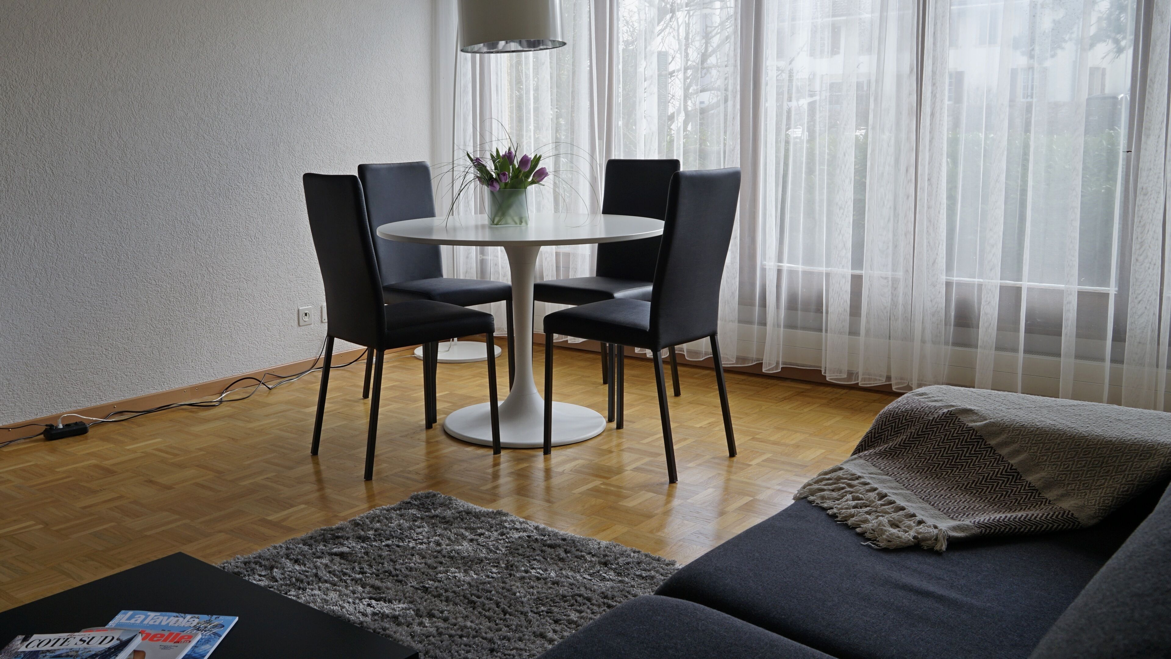 Una sala moderna con una mesa redonda de comedor, cuatro sillas negras, un sofá gris, un manto mullido gris y una ventana con cortinas translúcidas.