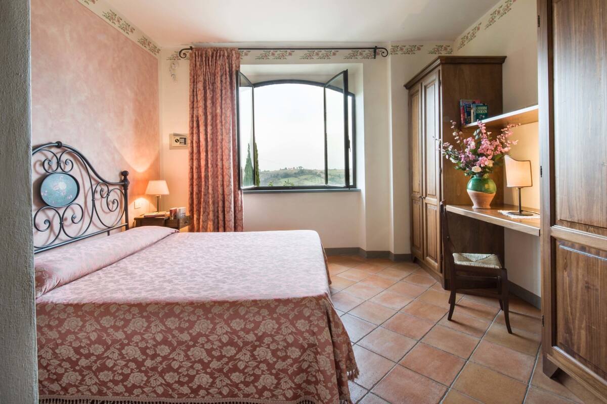 A bedroom with a bed, a window with curtains, a desk with a lamp, and a cabinet.