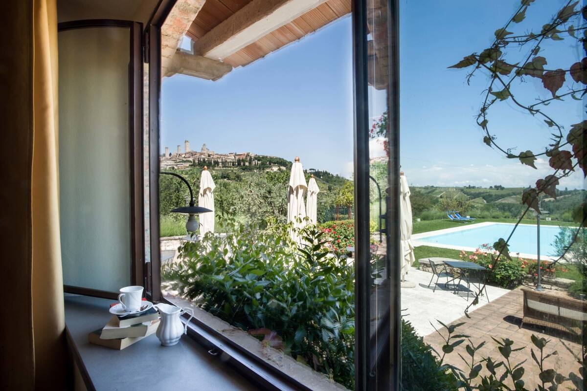 A view from a window overlooking a pool, greenery, and a distant building.