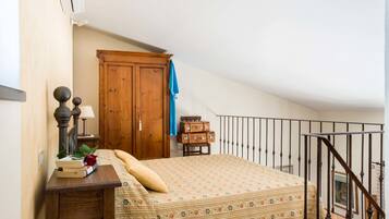 Un dormitorio con cama, un armario de madera, una pequeña mesa con libros y una rosa, y una escalera con barandilla de metal.