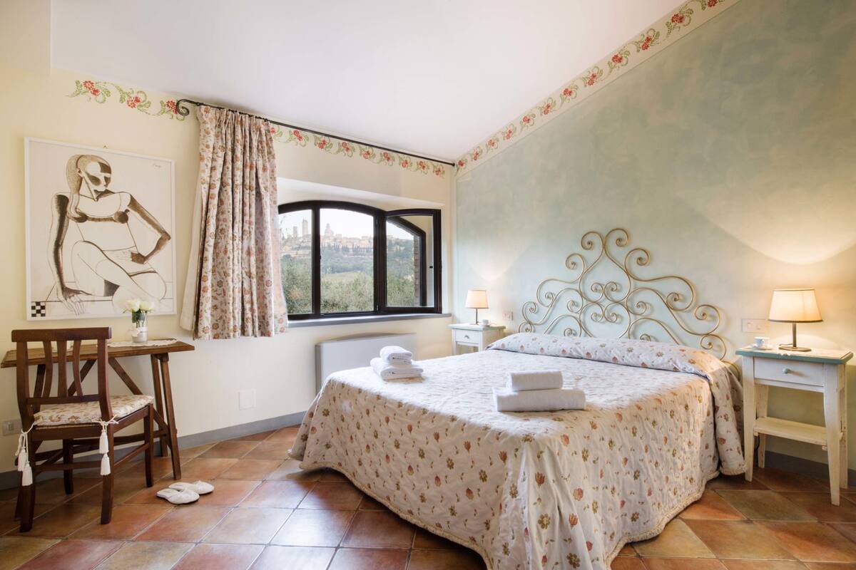 A bedroom with a bed, a desk, a chair, a window with a view, and a decorative headboard.