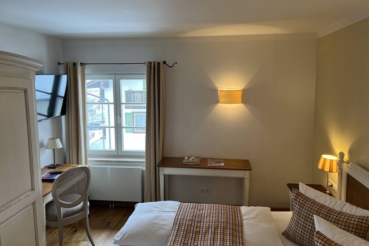 Ein Hotelzimmer mit einem Bett, einem Schreibtisch mit Stuhl, einem an der Wand befestigten Fernseher, einem Fenster mit Vorhängen und einer Wandlampe.