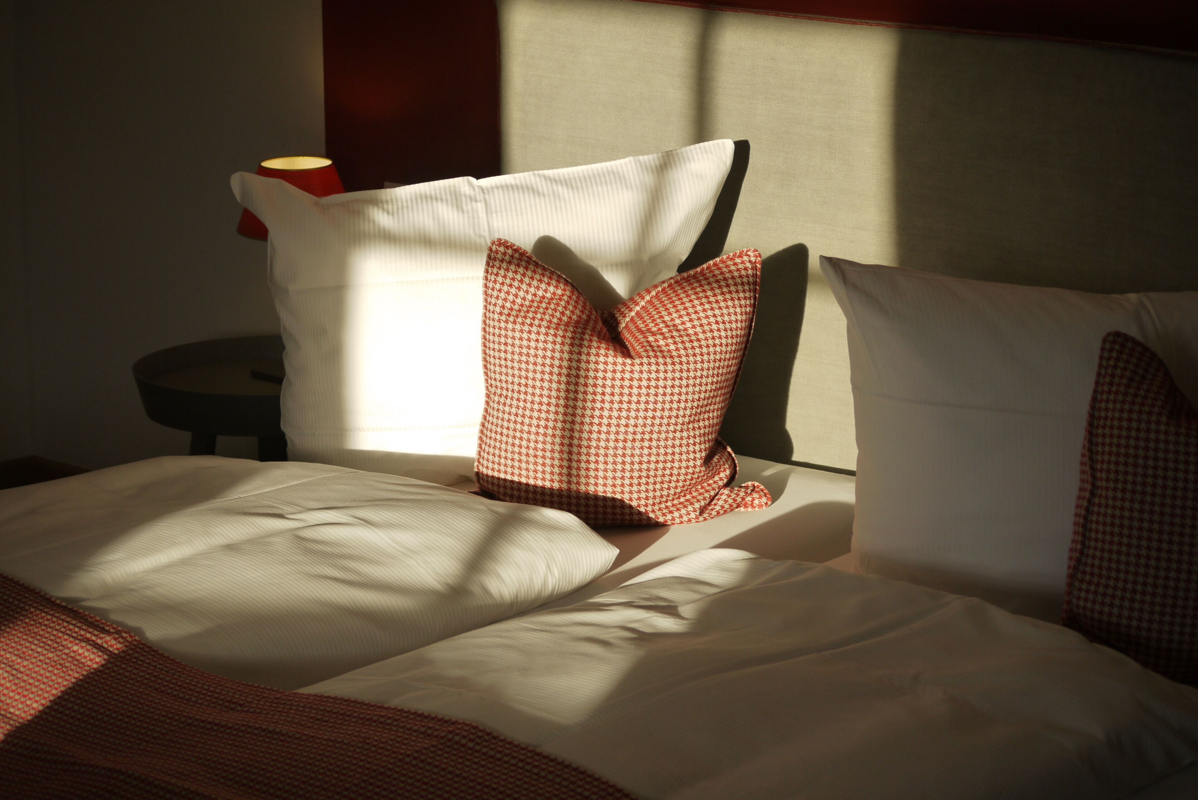 A neatly made bed with white and red patterned pillows, a red lamp on a bedside table, and a window casting shadows on the bed.