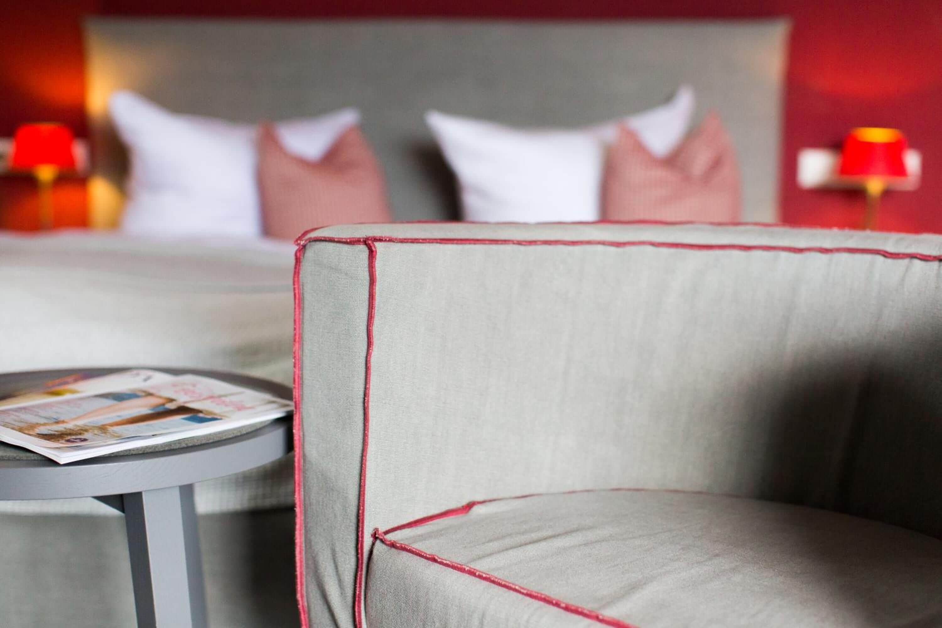 Een hotelkamer met een bed, kussens, een ronde tafel met een tijdschrift, en een stoel met rode stiksels.