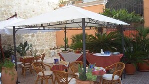 An outdoor patio with wicker chairs and tables, covered by a white canopy, surrounded by potted plants and a stone wall. - Sikania Suite (Pozzallo)
