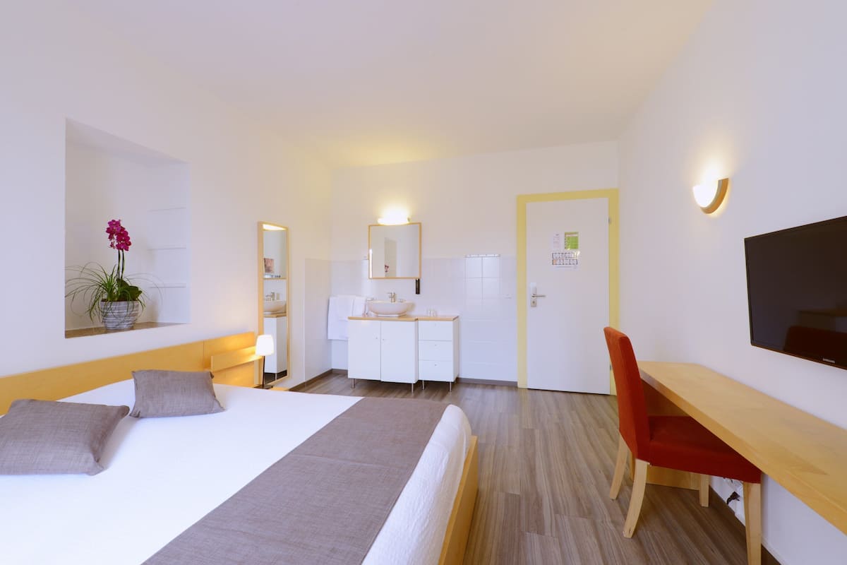A hotel room with a bed, bedside table, desk, chair, and a small plant.