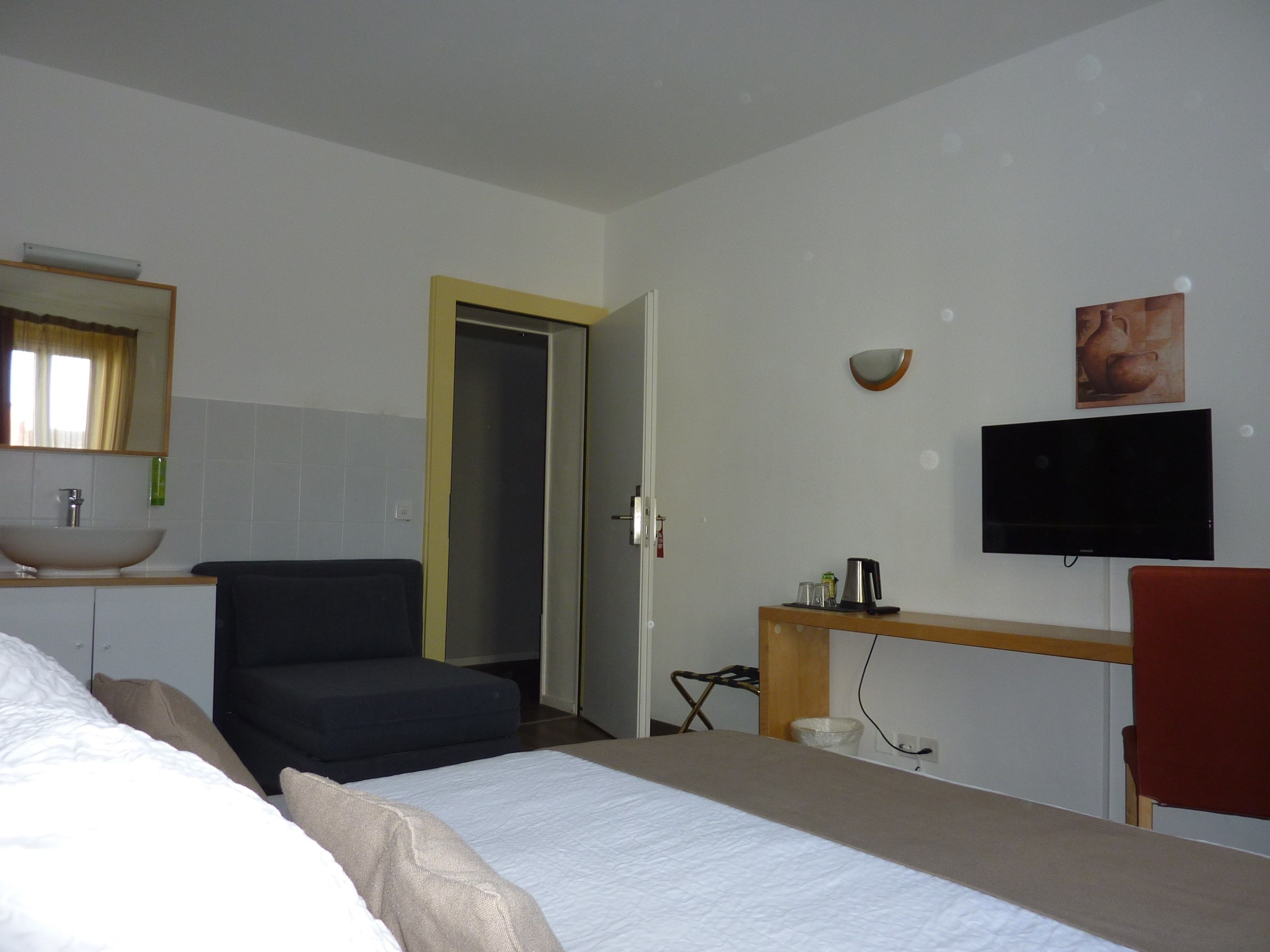 Ein Hotelzimmer mit Bett, Stuhl, Schreibtisch mit Flachbildfernseher, Spüle und einer Tür zu einem anderen Raum.