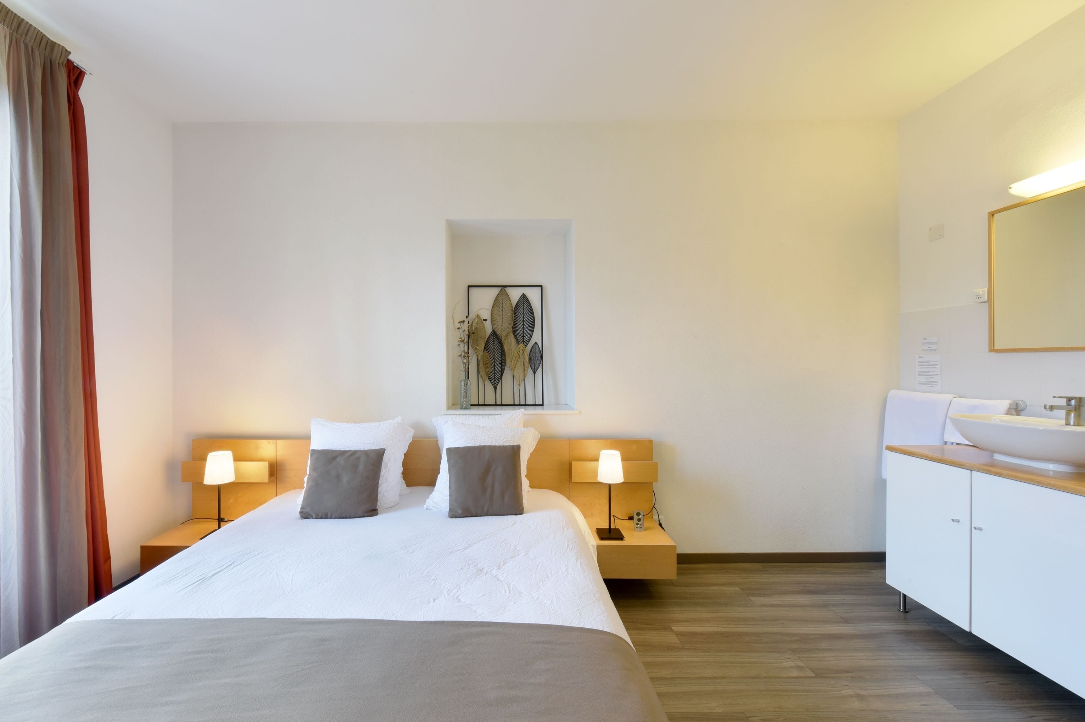 Ein Hotelzimmer mit einem großen Bett, Nachttischlampen, einem kleinen Kunstwerk an der Wand, einem Spiegel und einem Badezimmer mit Waschbecken und Schrank.