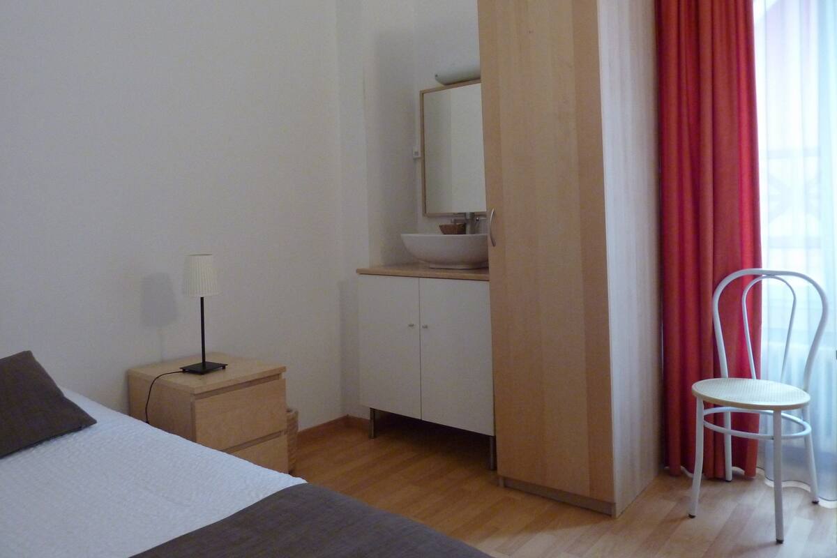 A bedroom with a bed, a bedside table with a lamp, a wardrobe, a mirror, a sink, and a chair.