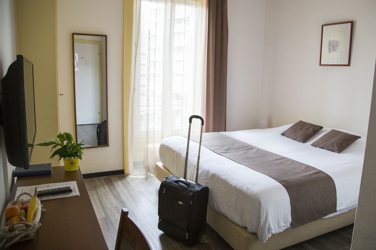 A hotel room with a bed, a desk with a plant, a suitcase, and a window with curtains.