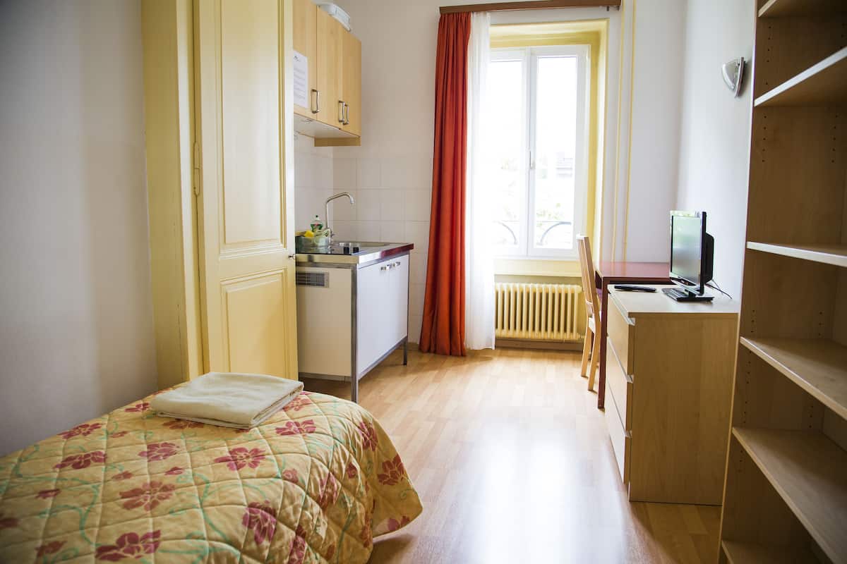 A small, well-lit room with a bed, a kitchenette, a desk, and a radiator.