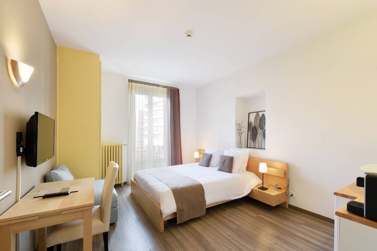 A hotel room with a bed, bedside tables, a desk, a chair, a television, and a window with curtains.