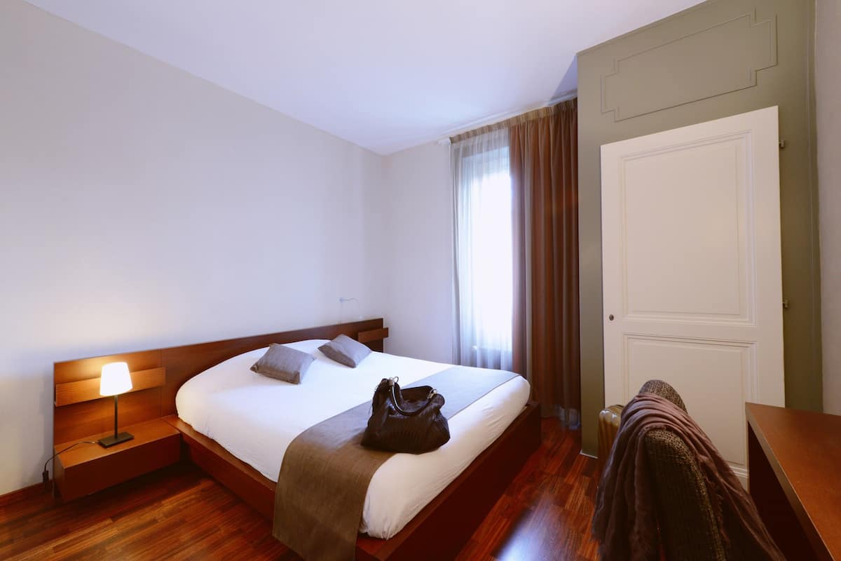 A neatly made bed with a brown bag on it, a bedside lamp, and a view of a window with curtains.