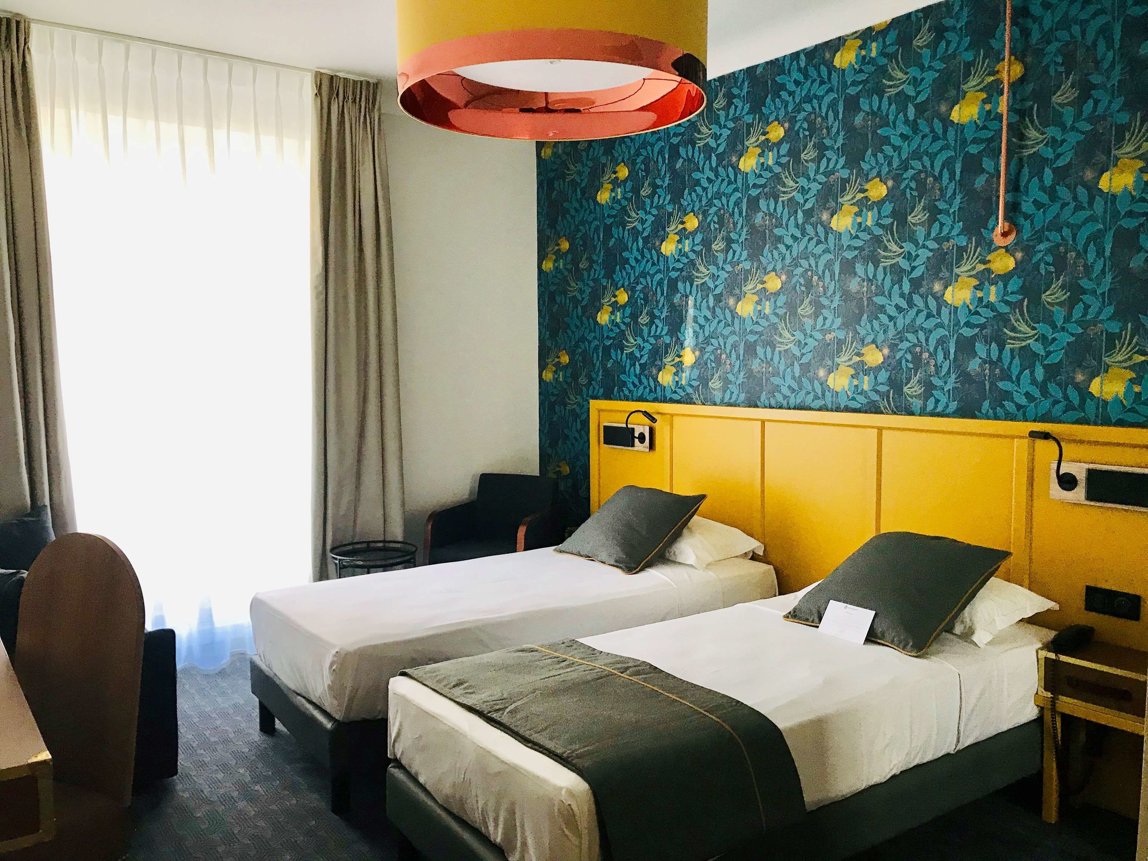Habitación de hotel con dos camas, un escritorio, una silla, una lámpara y una pared con estampado floral.