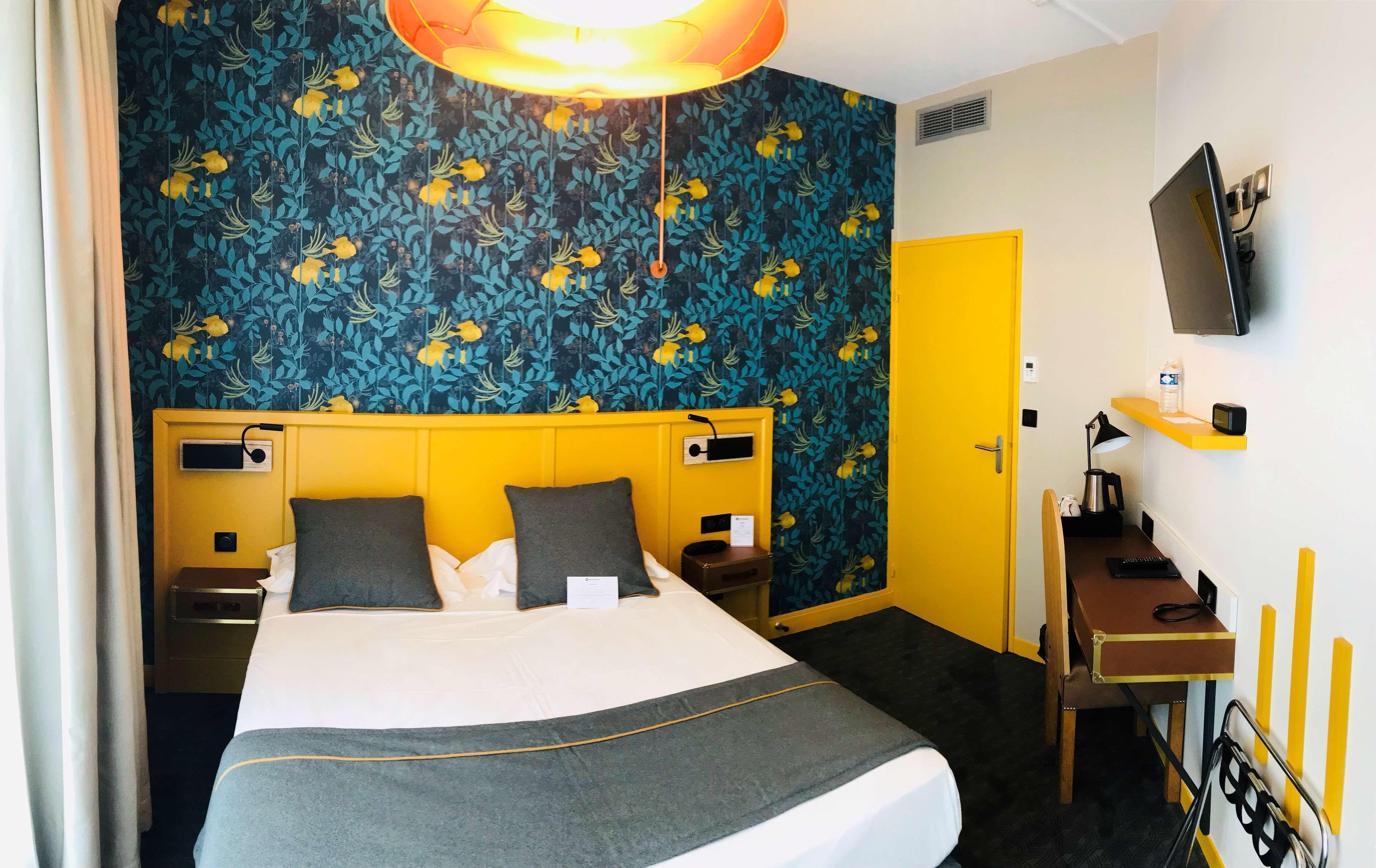 A hotel room with a bed, bedside tables, a desk with a chair, a TV mounted on the wall, and a door.