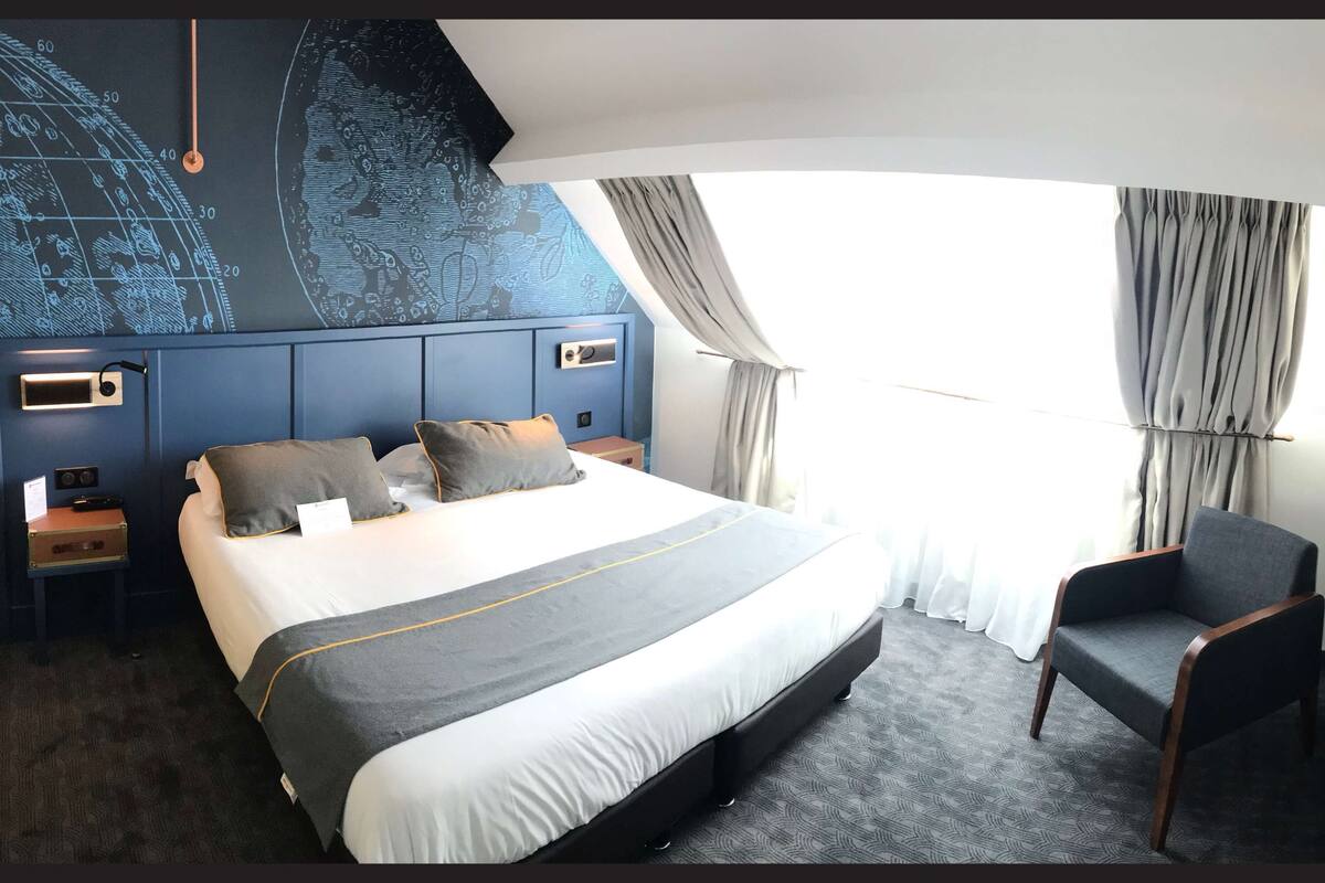 A hotel room with a large bed, a chair, a nightstand, a wall clock, and a window with curtains.