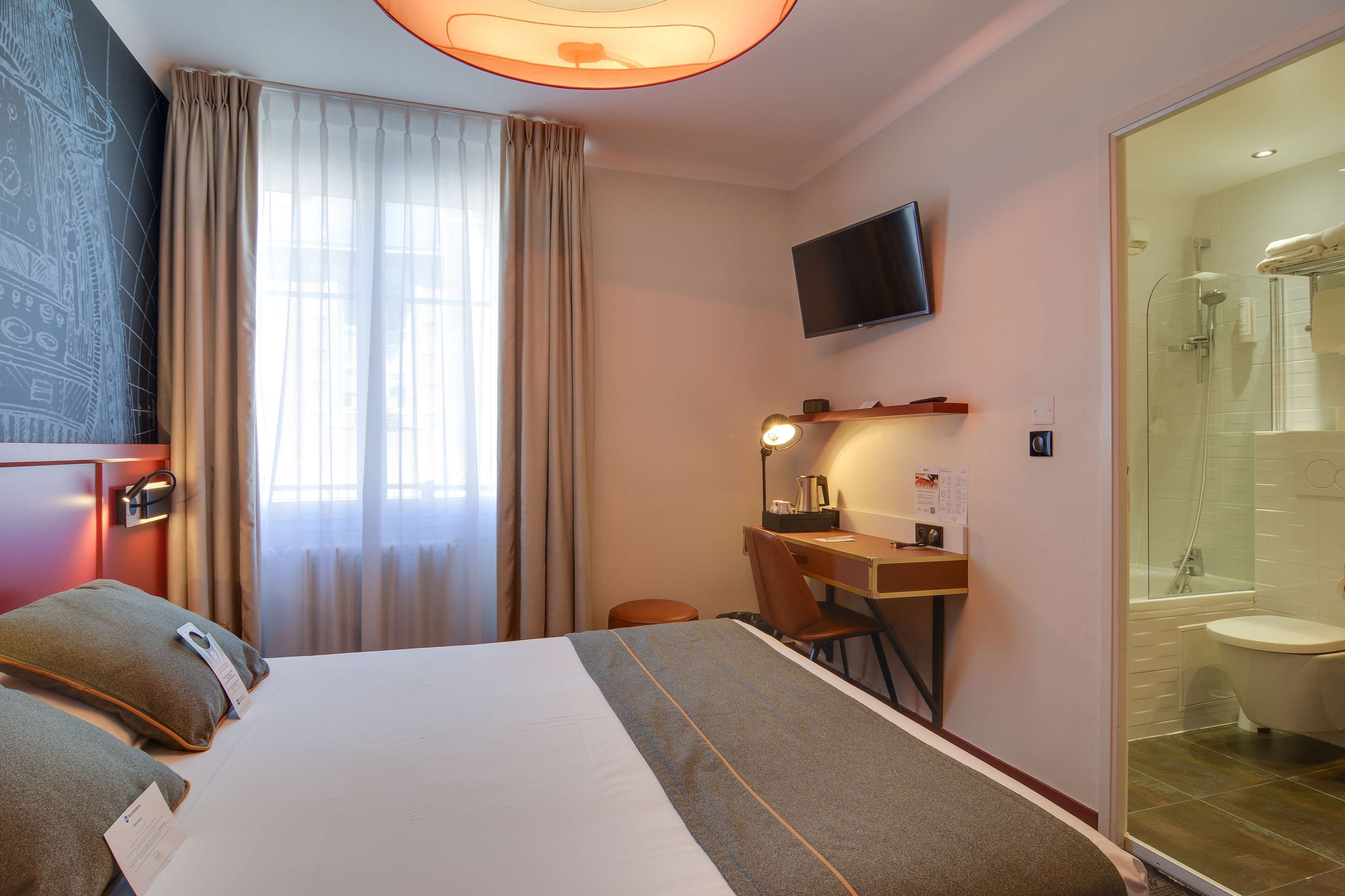A hotel room with a bed, desk, chair, TV, and a glass-enclosed shower area.