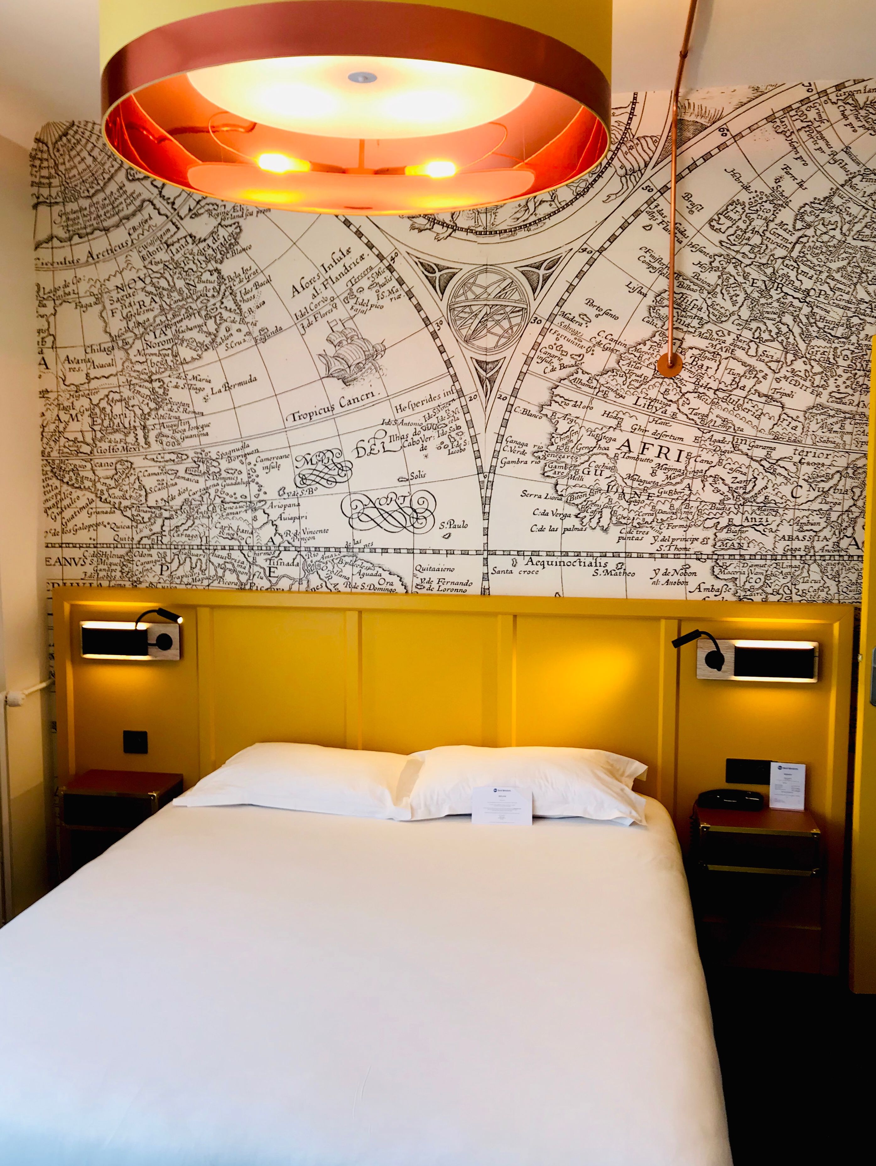A hotel room with a large bed, a map wall, and a modern ceiling light fixture.