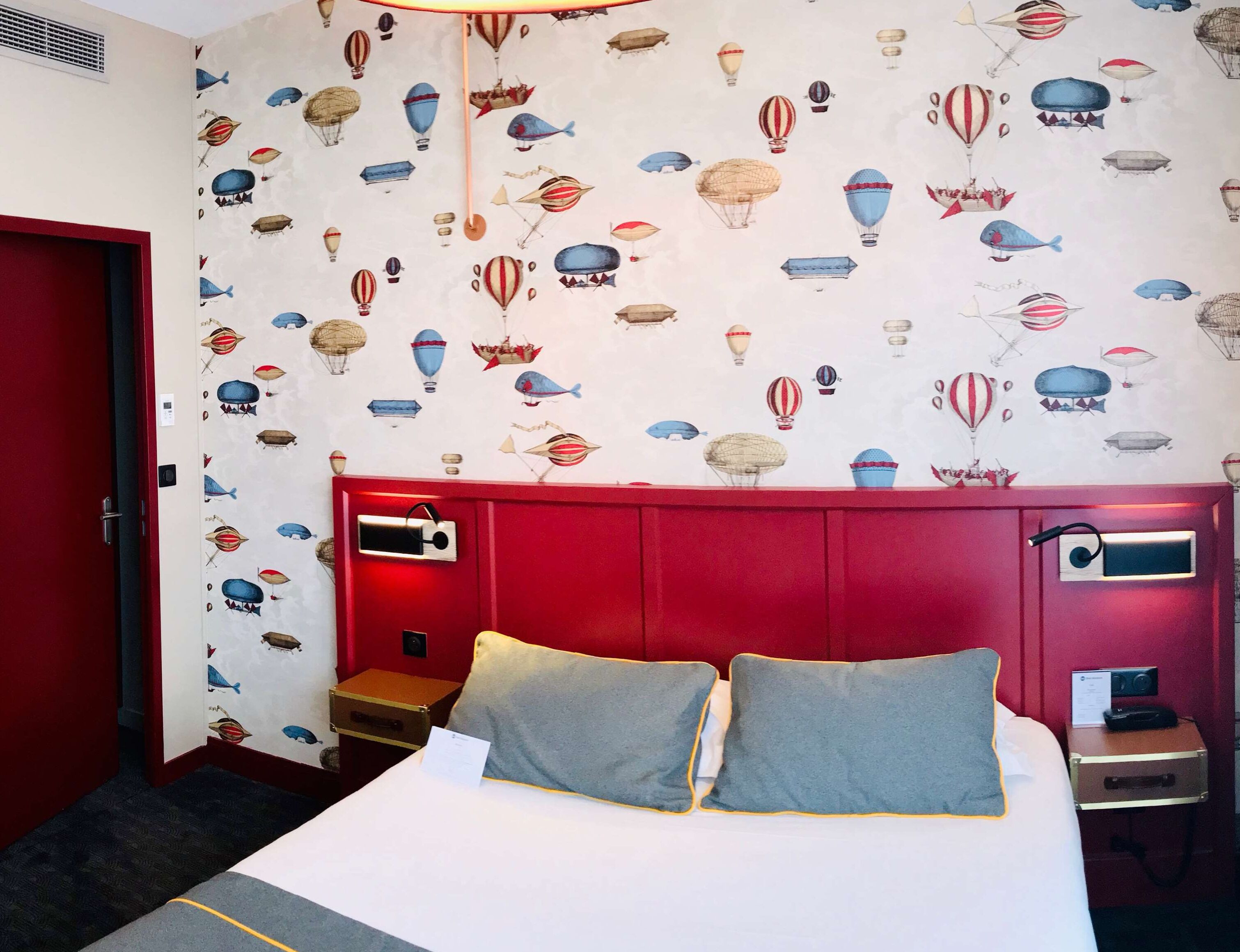 Quarto de hotel com cabeceira vermelha, cama com duas almofadas, mesa de cabeceira com abajur e parede decorada com padrão de balão de ar quente e peixes vintage.